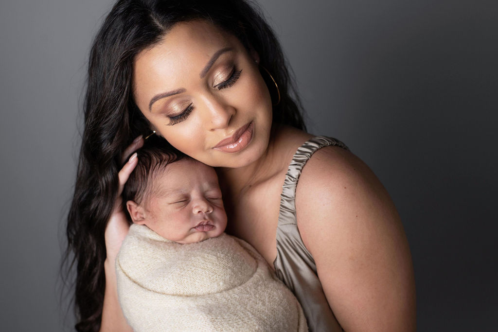 A mother in a champagne silk dress holding her swaddled newborn close to her cheek, eyes closed, against a soft grey backdrop.