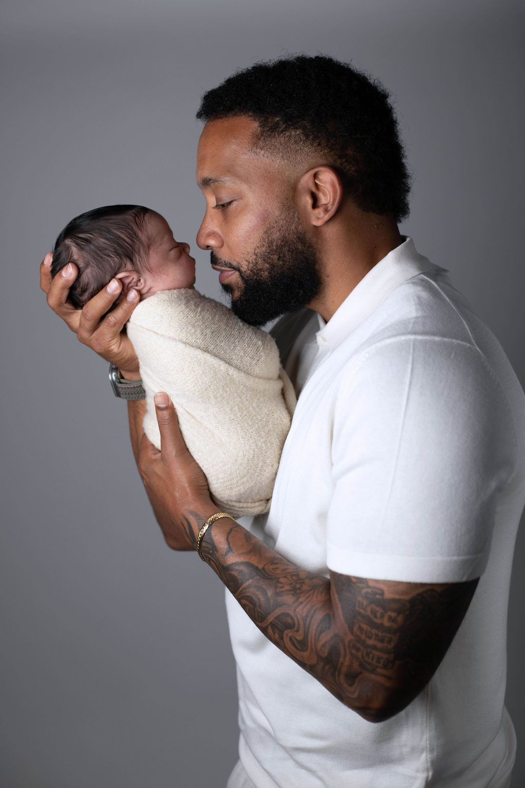 A father in a white polo shirt seen in profile, kissing the top of his swaddled newborn's head, holding the baby close to his face, against a soft grey backdrop.